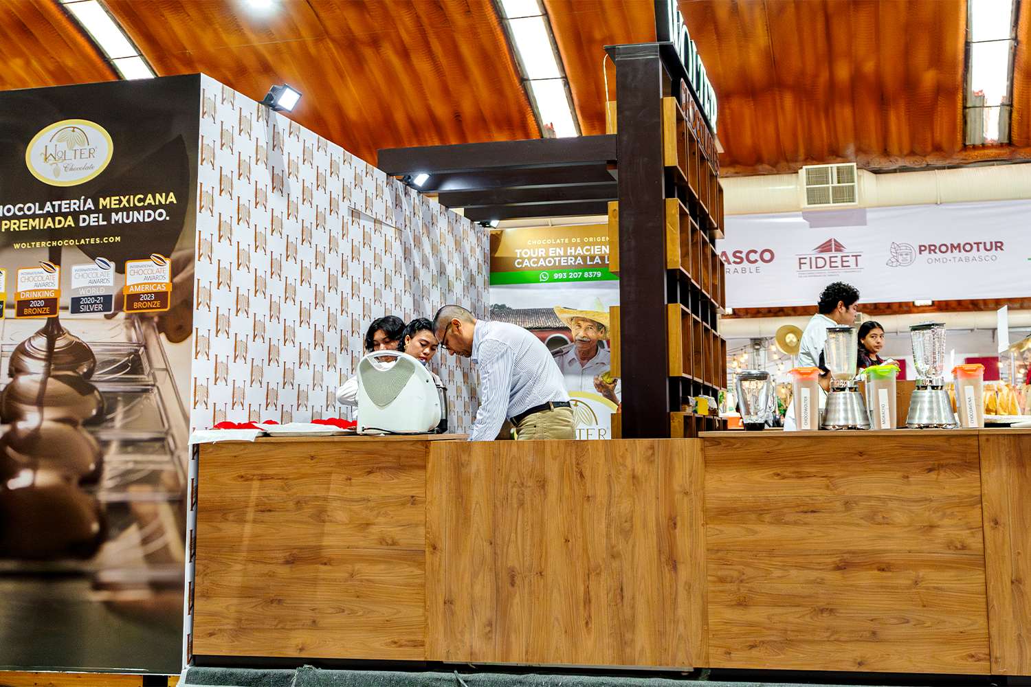 Vista lateral del stand de Wolter Chocolates mostrando el mostrador y la interacción con clientes.